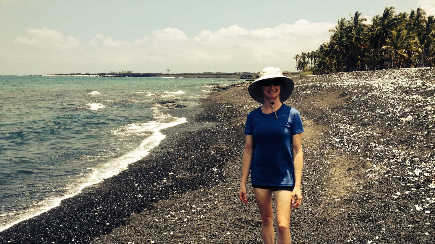 Carolyn Silan, PT, in a sun hat walking the black-sand shoreline on the Big Island, palms and surf behind her