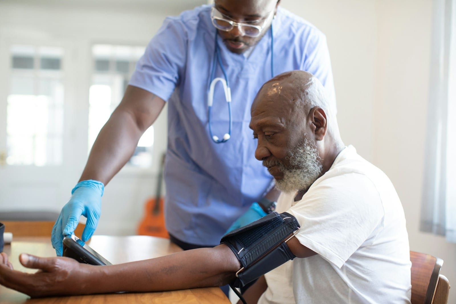 A nurse in blue scrubs takes an older man’s blood pressure with an arm cuff at a table