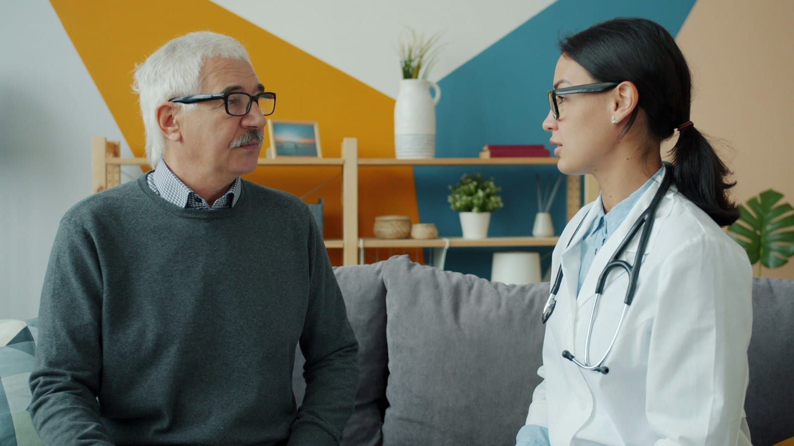 An older man with white hair and glasses talks with a doctor as they sit together on a sofa