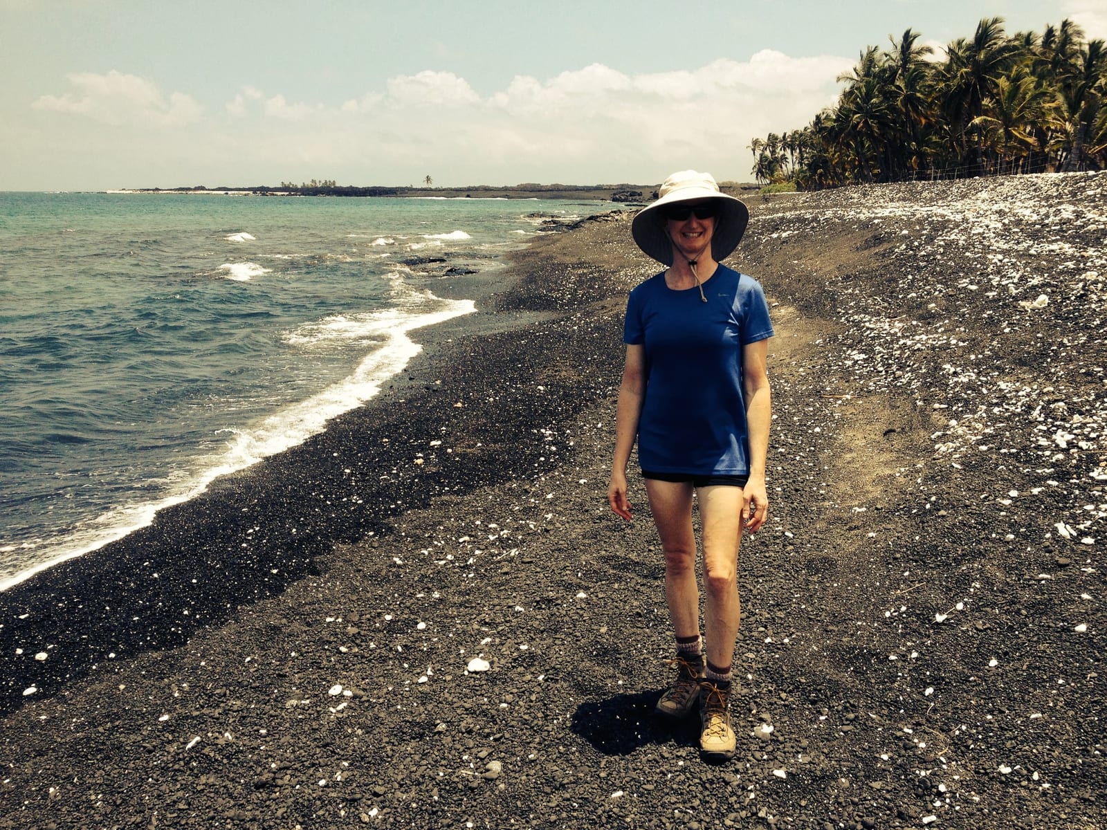 Carolyn Silan, PT, in a sun hat and sunglasses, standing on a black-sand beach with palm trees and the ocean behind her.