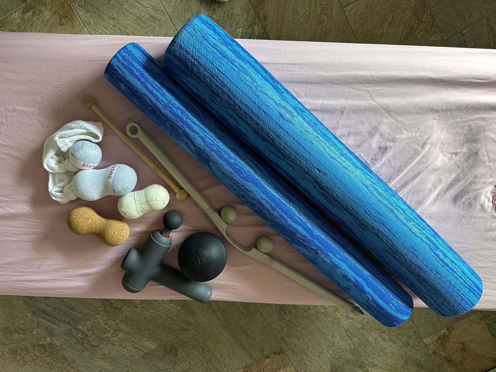 Self-massage tools laid out on a treatment table: two blue foam rollers, a wooden roller with knobs, a cork peanut ball, a firm black massage ball, a percussion massage gun, and tennis balls knotted inside socks.