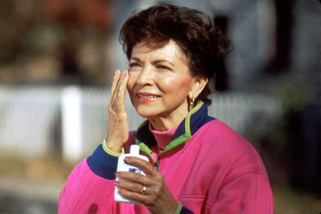 A woman in a pink jacket applying sunscreen to her face outdoors