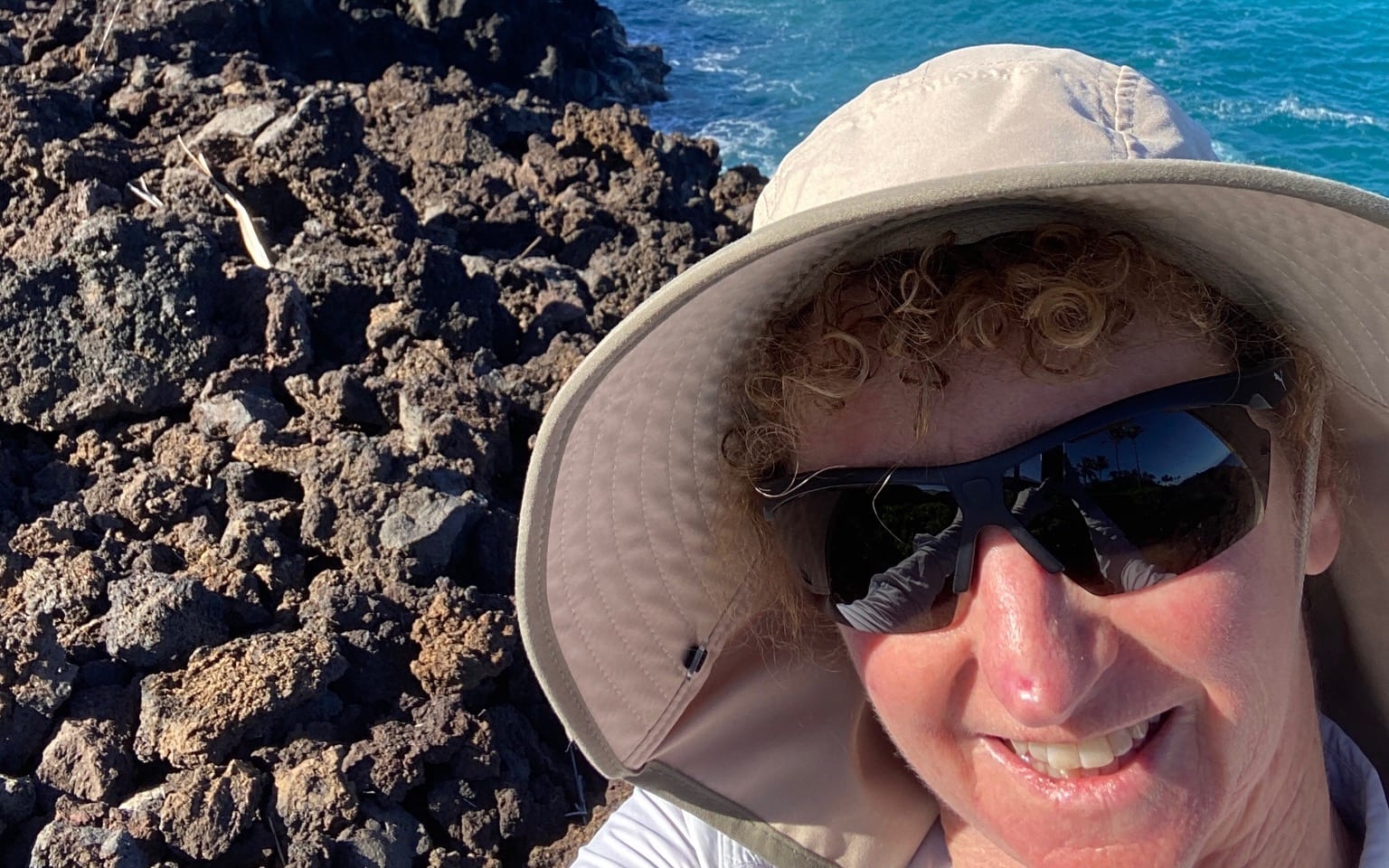 Carolyn in a wide-brimmed sun hat and sunglasses on the lava-rock coast, with the ocean behind her